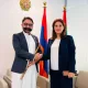 Two professionals shake hands in an office setting, with national flags and an emblem on the wall behind them.