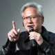 Man with gray hair and glasses speaks into a handheld microphone, raising his index finger to emphasize a point.
