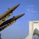 Two large missiles on display in front of a tall, arched monument against a blue sky.