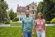 Smiling couple holding hands while walking across a manicured lawn in front of a brick house with a glass conservatory behind them.