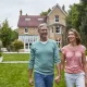 Smiling couple holding hands while walking across a manicured lawn in front of a brick house with a glass conservatory behind them.