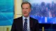Male news anchor in a dark suit and striped tie speaks from a television studio.