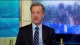 Male news anchor in a dark suit and striped tie speaks from a television studio.