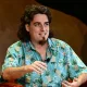 Man with shoulder-length dark hair and a goatee wearing a turquoise pineapple-patterned shirt, speaking on a stage with a microphone clipped to his collar.