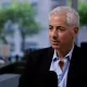 Middle‑aged man with short white hair wearing a dark blazer and light blue shirt speaks into a microphone in a bright office interview setting.