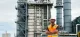 Construction worker in a bright orange safety vest and white hard hat standing with arms crossed in an industrial facility district.|A male worker at an oil refinery wearing a high-visibility vest and helmet, surveying the site.