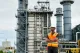 Construction worker in a bright orange safety vest and white hard hat standing with arms crossed in an industrial facility district.|A male worker at an oil refinery wearing a high-visibility vest and helmet, surveying the site.