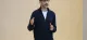 Man wearing glasses and a navy zip sweater speaks, gesturing with his hands in front of a beige wall.