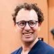 Smiling man with curly dark hair and blue round glasses, wearing a navy sweater, against an orange background:** portrait-style image