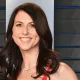 Smiling woman with long dark hair in a red dress and pearl necklace, posing against a dark backdrop with blue accents.